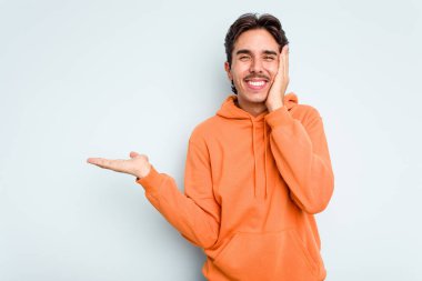 Mavi arka planda izole edilmiş genç İspanyol bir adam avucunun içinde fotokopi alanı tutuyor, el üstünde tutuyor. Şaşırmış ve mutlu..