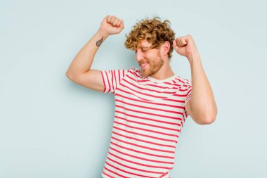 Mavi arka planda izole genç beyaz adam özel bir günü kutluyor, atlıyor ve enerjiyle kollarını kaldırıyor..