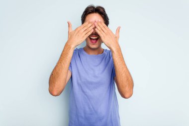 Mavi arka planda izole edilmiş genç İspanyol adam elleriyle gözlerini kapatıyor, geniş bir gülümsemeyle bir sürpriz bekliyor..