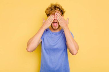 Sarı arka planda izole edilmiş genç beyaz adam elleriyle gözlerini kapatıyor, geniş bir gülümsemeyle bir sürpriz bekliyor..