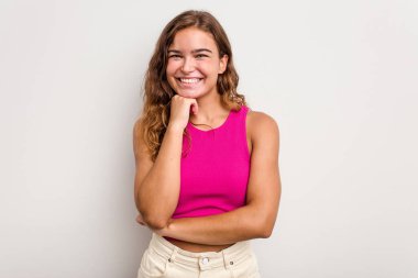 Mavi arka planda izole edilmiş genç beyaz kadın mutlu ve kendinden emin gülümsüyor, elleriyle çenesine dokunuyor..