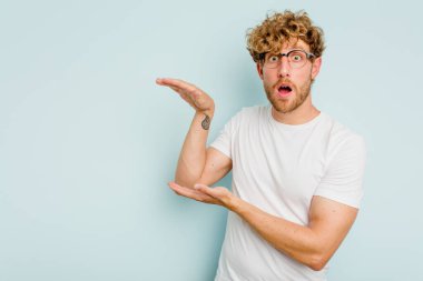 Mavi arka planda izole genç beyaz adam şok ve hayret içinde elleri arasında bir kopya boşluk tutuyor.