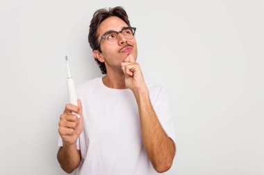 Beyaz arka planda elektrikli diş fırçası tutan İspanyol genç adam şüpheli ve şüpheci bir ifadeyle yana bakıyor..