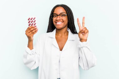 Genç Afrikalı Amerikalı eczacı kadın mavi arka planda iki numarayı parmaklarıyla gösteriyor..