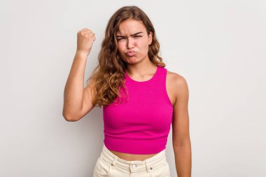 Mavi arka planda izole edilmiş genç beyaz kadın yumruğunu kameraya gösteriyor, agresif yüz ifadesi..