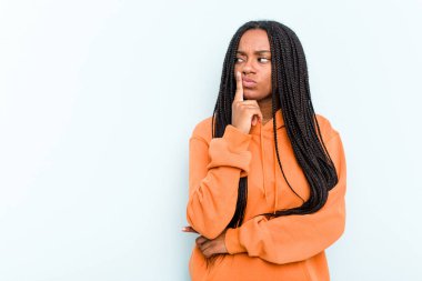 Mavi arka planda saç örgüsü olan genç Afrikalı Amerikalı kadın şüpheli ve şüpheci bir ifadeyle yana bakıyor..
