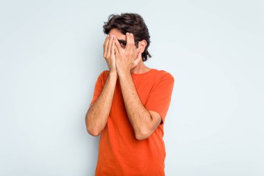 Mavi arka planda izole edilmiş genç İspanyol adam kameraya parmaklarıyla göz kırpıyor, yüzünü kapatmaktan utanıyor..