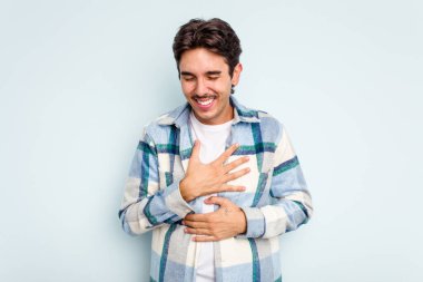 Mavi arka planda izole edilmiş genç İspanyol adam mutlu bir şekilde gülüyor ve ellerini karnının üstünde tutarak eğleniyor..