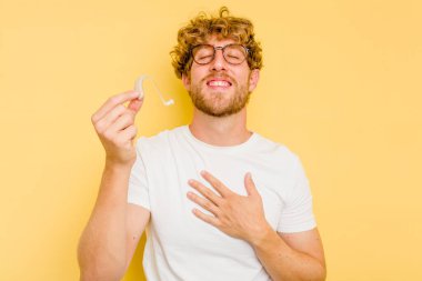 Sarı arka planda izole edilmiş işitme cihazı takan genç beyaz bir adam elini göğsünde tutarak kahkahalar atıyor..