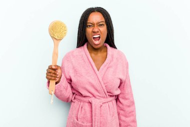 Elinde küvet fırçası tutan genç Afrikalı bir kadın mavi arka planda izole edilmiş çok kızgın ve agresif çığlıklar atıyor..