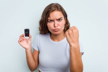 Young caucasian woman holding car keys isolated on blue background showing fist to camera, aggressive facial expression.