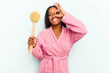 Genç Afrikalı Amerikalı bir kadın elinde bir küvet fırçası tutarken mavi arka planda izole edilmiş..