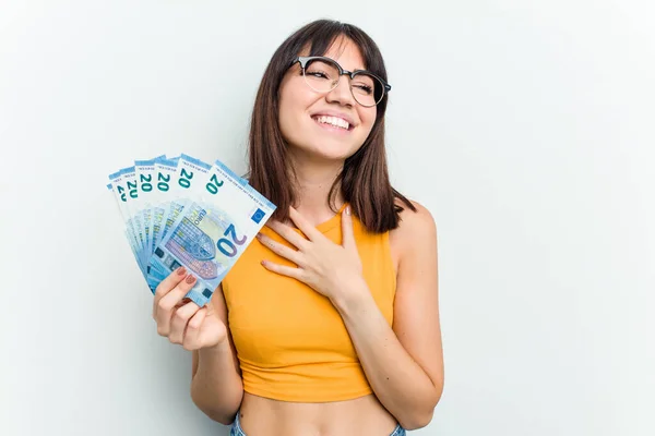 Young caucasian woman holding a banknotes isolated on blue background laughs out loudly keeping hand on chest.