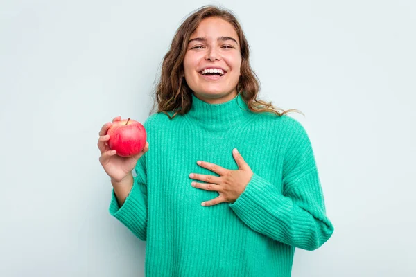 年轻的高加索女人 有一个蓝色背景的苹果 大声笑着把手放在胸前 — 图库照片