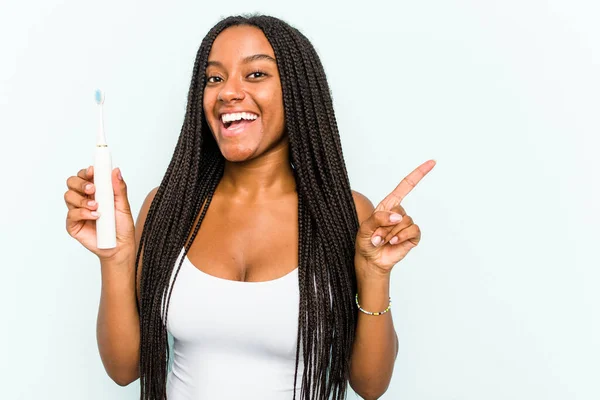 年轻的非洲裔美国女人拿着被蓝色背景隔离的电动牙刷 微笑着指着旁边 在空白的地方展示着一些东西 — 图库照片
