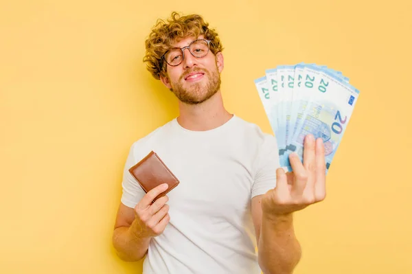 Young caucasian man holding banknotes and wallet isolated on yellow background