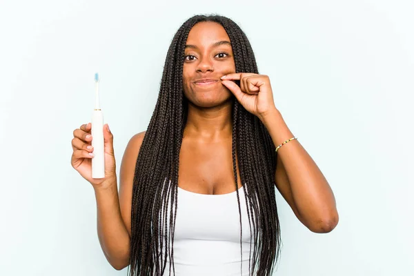 若いですアフリカ系アメリカ人女性を保持しています電動歯ブラシ隔離上の青の背景に指で唇に秘密を保持 — ストック写真