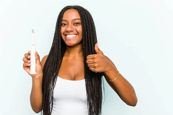年轻的非裔美国女人拿着被蓝色背景隔离的电动牙刷 微笑着举起大拇指 — 图库照片