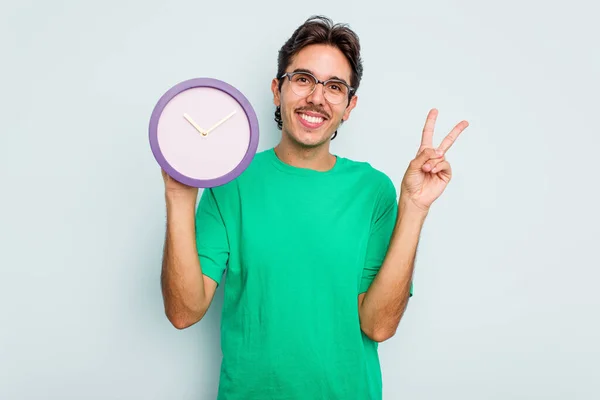 白い背景に時計を持つ若いヒスパニック系の男喜びと屈託のない指で平和のシンボルを示す — ストック写真