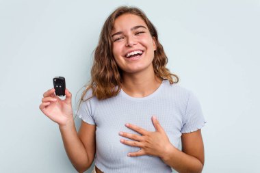 Young caucasian woman holding car keys isolated on blue background laughs out loudly keeping hand on chest.