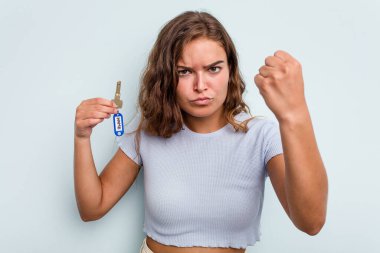 Young caucasian woman holding home keys isolated on blue background showing fist to camera, aggressive facial expression.