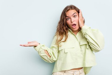 Mavi arka planda izole edilmiş genç beyaz kadın, avuç içinin fotokopi alanını tutmaktan etkilenmiş..