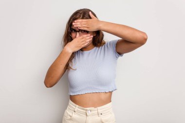 Mavi arka planda izole edilmiş genç beyaz kadın kameraya parmaklarıyla göz kırpıyor, yüzünü kapatmaktan utanıyor..