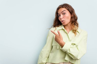 Mavi arka planda izole edilmiş genç beyaz kadın gülümsüyor ve boş alanda bir şeyler gösteriyor..