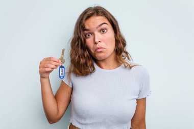 Young caucasian woman holding home keys isolated on blue background shrugs shoulders and open eyes confused.