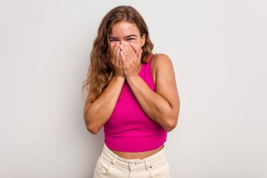 Mavi arka planda izole edilmiş genç beyaz kadın bir şeye gülüyor, elleriyle ağzını kapatıyor..