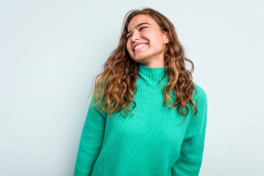 Genç beyaz kadın mavi arka planda izole edilmiş rahat ve mutlu bir şekilde gülüyor, boynunda dişlerini gösteriyor..