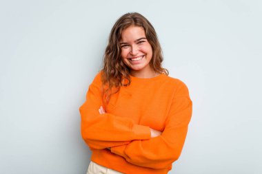 Mavi arka planda izole edilmiş genç beyaz kadın kendini güvende hissediyor, kararlılıkla kollarını kavuşturuyor..