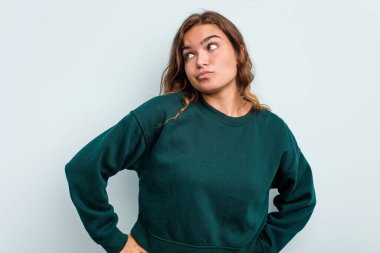 Kafkasyalı genç bir kadın mavi arka planda izole edilmiş, kafası karışmış, şüpheli ve kendinden emin değil..