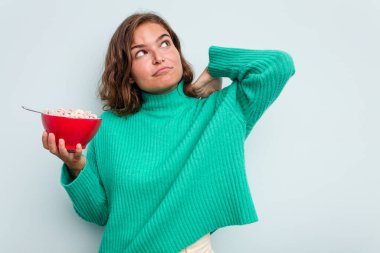 Beyaz bir kadın elinde bir kase mısır gevreği tutarken mavi arka planda izole edilmiş kafasının arkasına dokunuyor, düşünüyor ve bir seçim yapıyor..