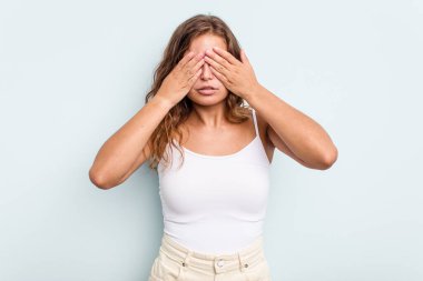 Mavi arka planda izole edilmiş genç beyaz kadın elleriyle gözlerini kapatmaktan korkuyor..