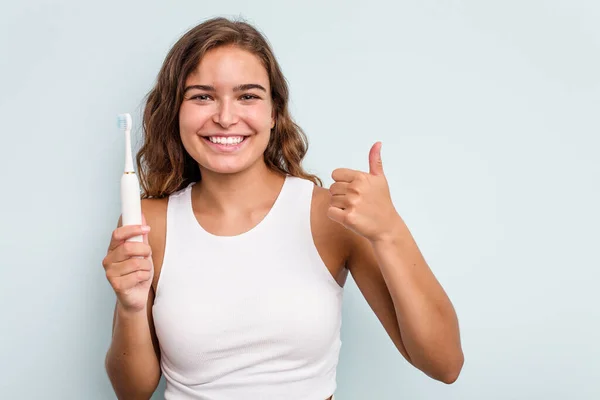 年轻的高加索女人拿着电动牙刷孤立的蓝色背景微笑着举起大拇指 — 图库照片