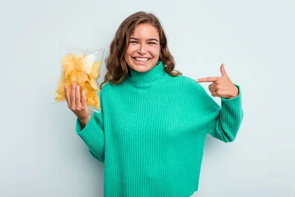 年轻的高加索女人拿着土豆条 与蓝色背景的人隔离 手指着衬衫的复制品空间 自豪而自信 — 图库照片