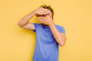 Sarı arka planda izole edilmiş genç beyaz adam kameraya parmaklarıyla göz kırpıyor, yüzünü kapatmaktan utanıyor..
