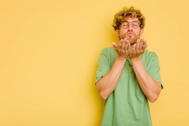 Sarı arka planda izole edilmiş genç, beyaz bir adam. Dudaklarını kıvırıyor, avuç içlerini tutuyor ve hava öpücüğü yolluyor..