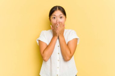 Sarı arka planda izole edilmiş genç Asyalı kadın şok olmuş, ağzı ellerle kaplanmış, yeni bir şey keşfetmek için sabırsızlanıyor..