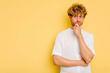 Sarı arka planda izole edilmiş genç beyaz adam şüpheli ve şüpheci bir ifadeyle yana bakıyor..