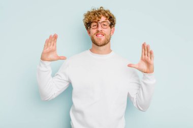 Kafkasyalı genç adam mavi arka planda izole edilmiş. Elinde avuç içi olan bir şey var ve kameraya sunuyor..