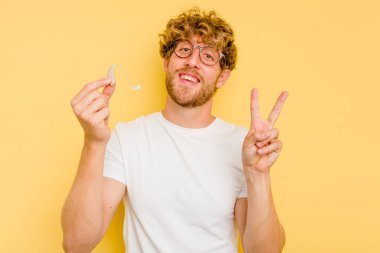 İşitme cihazı takan genç beyaz adam sarı arka planda izole edilmiş neşeli ve kaygısız parmaklarıyla barış sembolü gösteriyor..