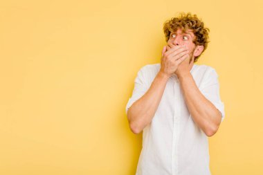 Kafkasyalı genç adam sarı arka planda izole edilmiş. Düşünceli bir şekilde, ağzını eliyle kapatan bir fotokopi odasına bakıyor..