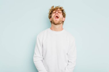 Kafkasyalı genç adam mavi arka planda izole edilmiş rahat ve mutlu bir şekilde gülüyor, boynunda dişlerini gösteriyor..
