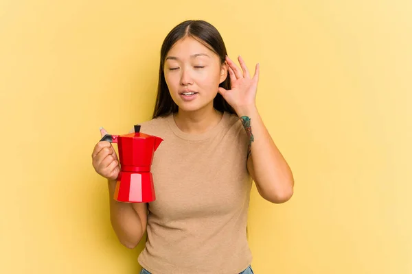 若いアジアの女性がコーヒーメーカーを保持している黄色の背景に隔離されたゴシップを聞いてみる — ストック写真