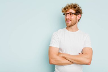 Mavi arka planda izole edilmiş genç beyaz adam kollarını kavuşturarak gülümsüyor..