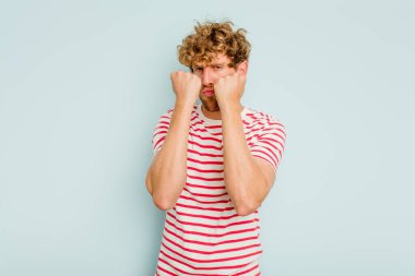 Kafkasyalı genç bir adam mavi arka planda izole edilmiş. Yumruk atıyor, sinirleniyor, kavga ediyor, kavga ediyor..