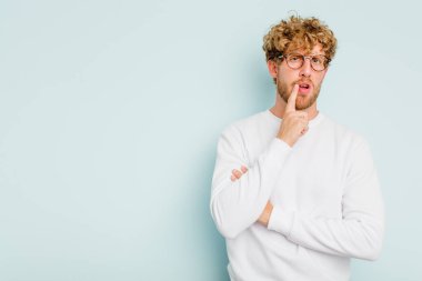 Mavi arka planda izole edilmiş genç beyaz adam şüpheli ve şüpheci bir ifadeyle yana bakıyor..