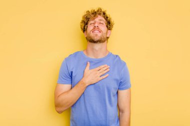 Sarı arka planda izole edilmiş genç beyaz adam yüksek sesle gülüyor ve elini göğsüne koyuyor..